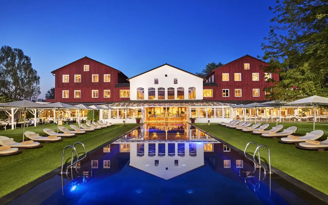 Abendansicht des Bleiche Resort & Spa mit beleuchtetem Hotelgebäude, Aussenpool und Liegewiesen unter blauem Himmel.
