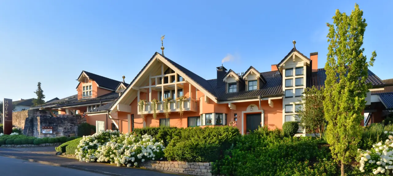 Aussenansicht des Landhaushotels Hof Wasserkuppe mit oranger Fassade, Balkon und blühenden Hortensien unter blauem Himmel.