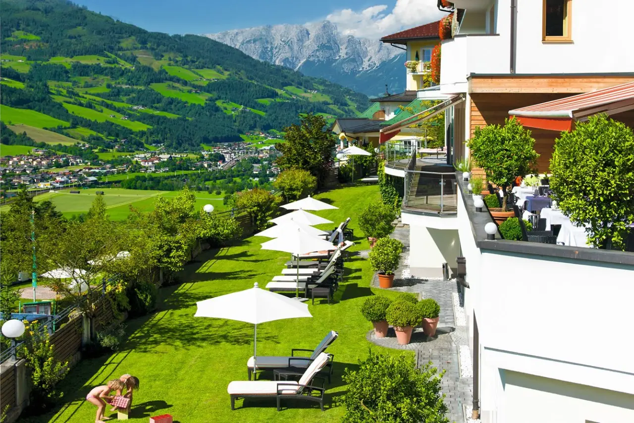 Panoramablick auf das Wellness Hotel Zinnkrügl mit sonniger Liegewiese, Restaurantterrasse und Alpenbergwelt im Hintergrund.