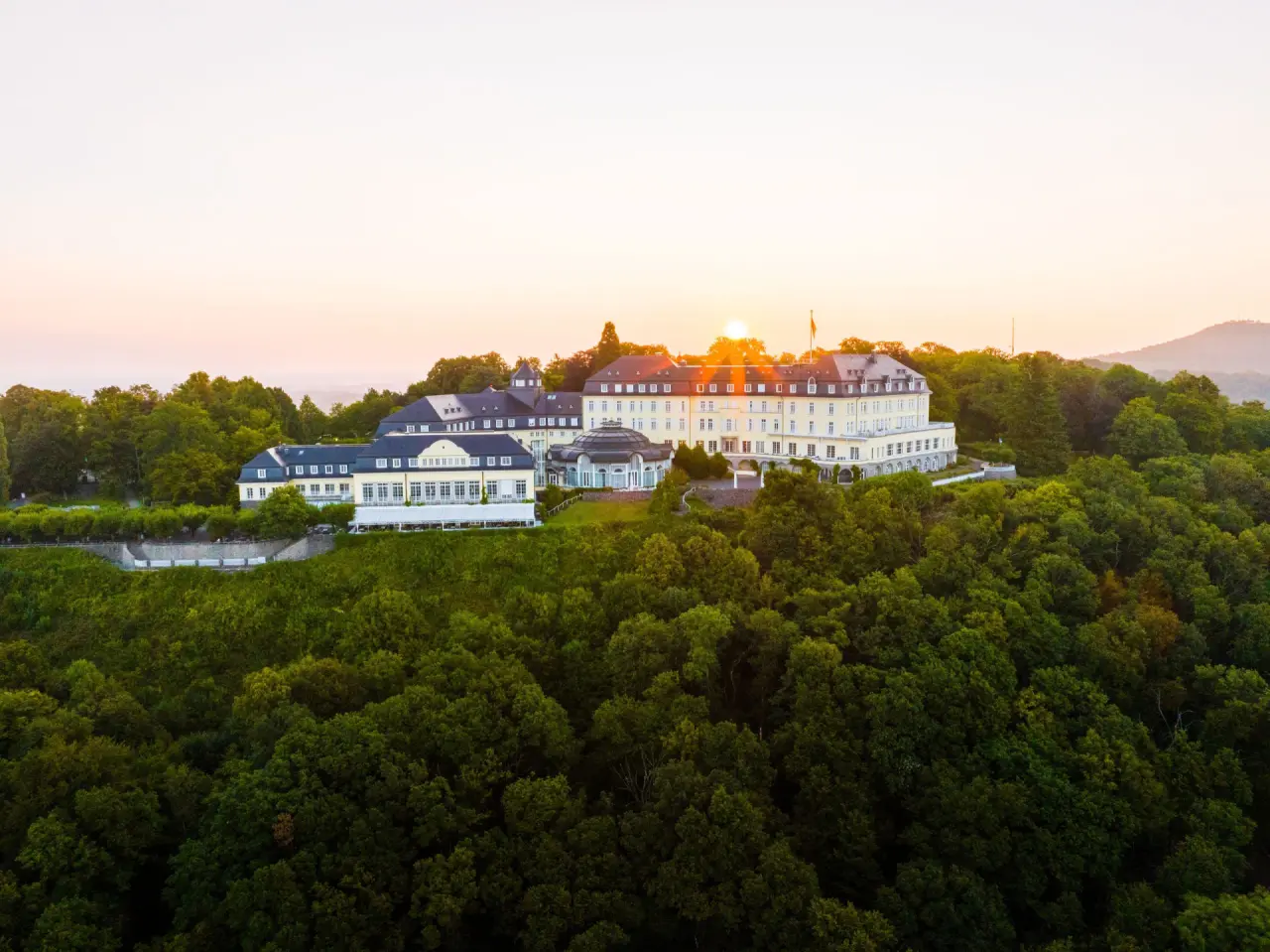 Majestätisches Steigenberger Grand Petersberg Hotel bei Sonnenuntergang, umgeben von grünen Wäldern.