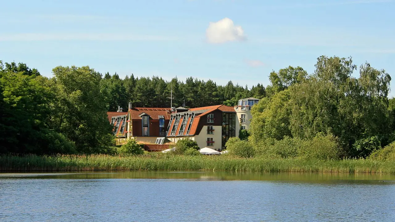 Aussenansicht des Hotel & Spa Sommerfeld am See, umgeben von grünen Bäumen und Schilf unter blauem Himmel.
