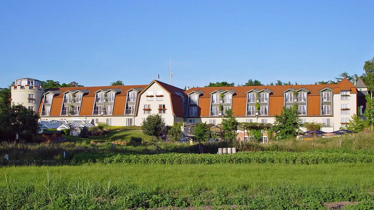 Aussenansicht des Hotel & Spa Sommerfeld mit rotem Dach und Balkonen, umgeben von Gruenflaechen unter blauem Himmel.