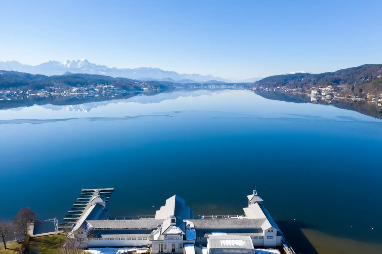 Luftaufnahme vom historischen Werzer-Bad am Wörthersee mit spiegelnder Wasseroberfläche und Alpenpanorama, Werzers Hotel Resort Pörtschach.