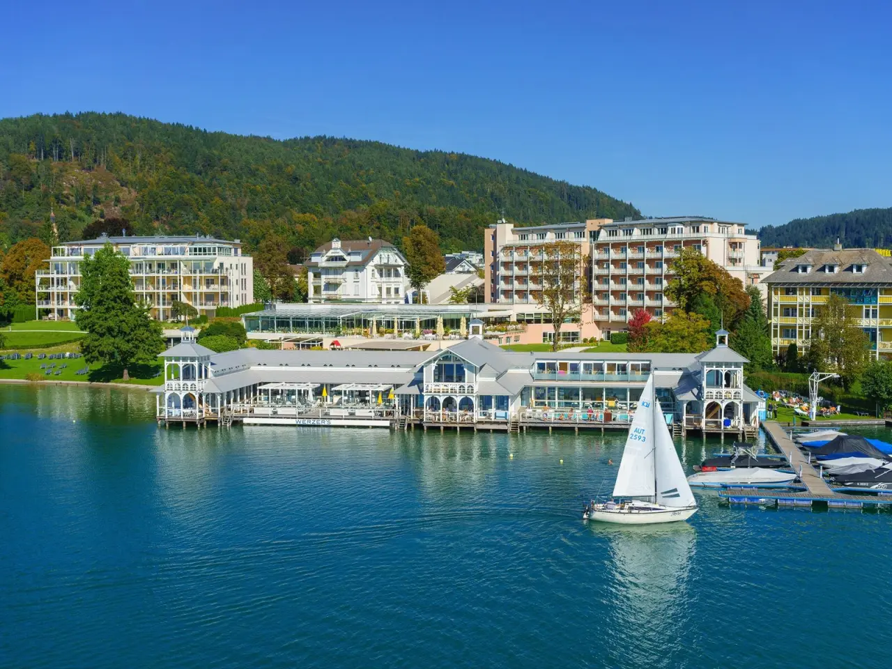 Luftaufnahme des Werzers Hotel Resort Pörtschach am Wörthersee mit historischem Badehaus, Segelboot und bewaldeten Hügeln.