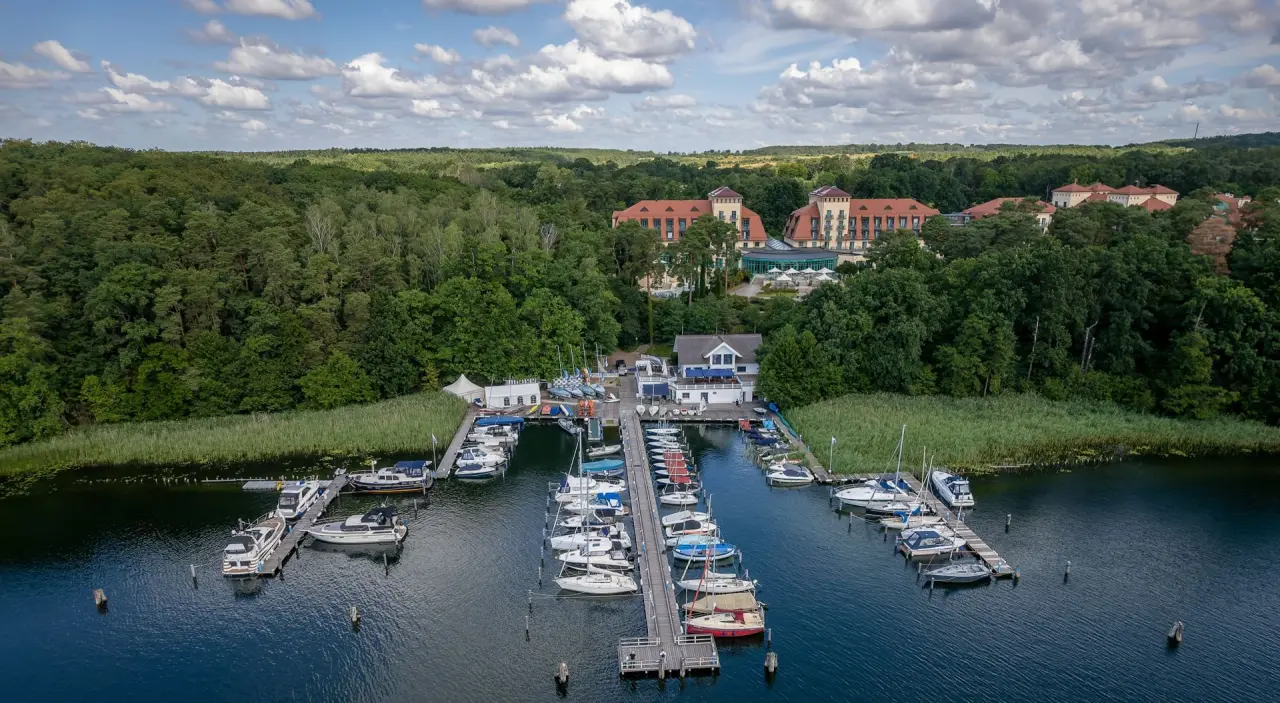 Luftaufnahme des Precise Resort Bad Saarow am Scharmützelsee mit Hotelgebäude, Wald und belebter Marina unter blauem Himmel.