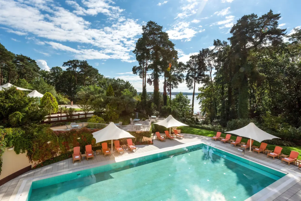 Aussenpool mit Sonnenliegen und Schirmen, umgeben von grüner Natur und Blick auf den Scharmützelsee im Precise Resort Bad Saarow.