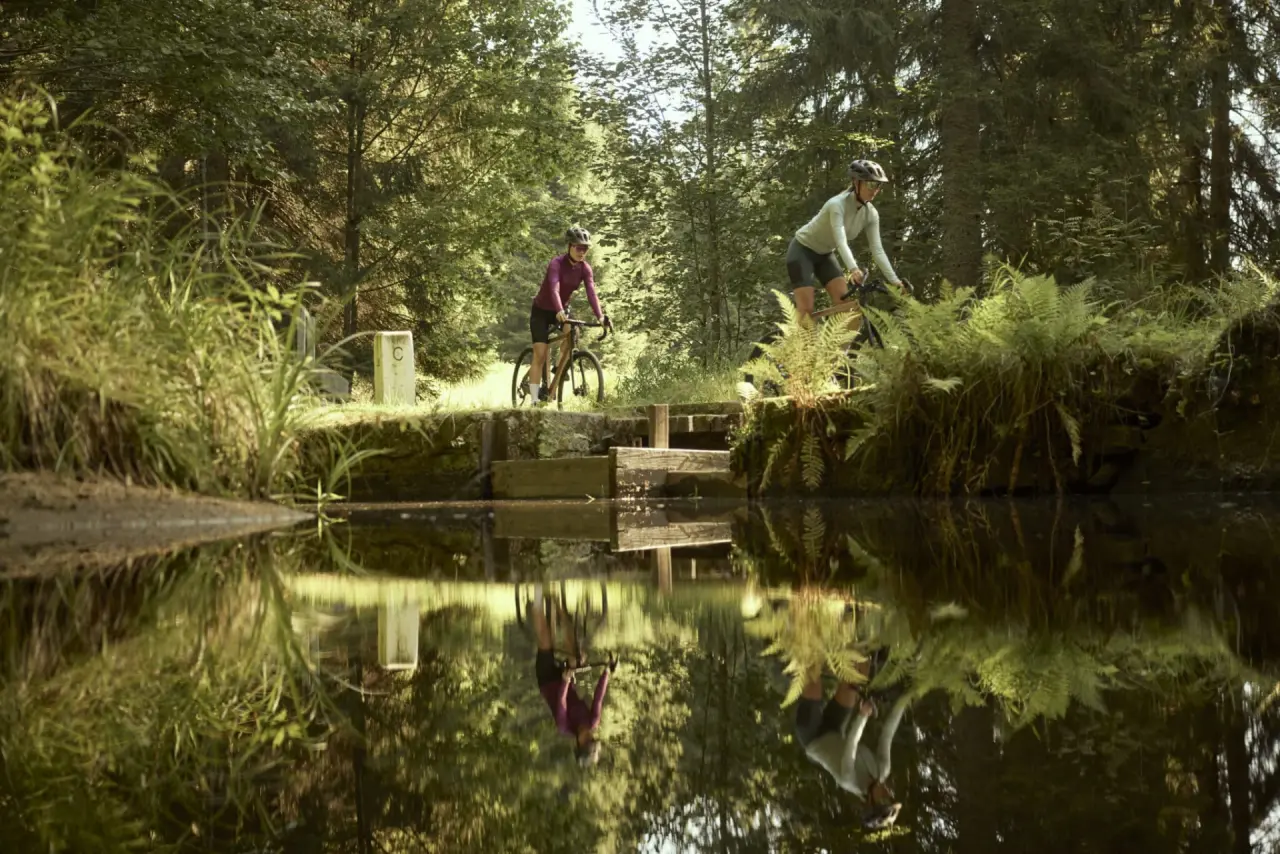 Zwei Radfahrerinnen auf Mountainbikes im Wald mit Spiegelung im Wasser, AVIVA****s make friends Hotel.