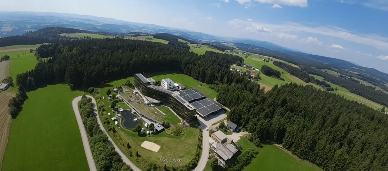 Luftaufnahme des AVIVA****s make friends Hotels eingebettet in grüne Hügellandschaft mit Naturteich und Wald.