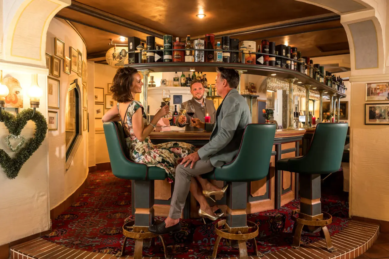 Gemütliche Hotelbar im Romantik Hotel Wastlwirt mit Gästen, die Wein genießen und mit dem Barkeeper plaudern.