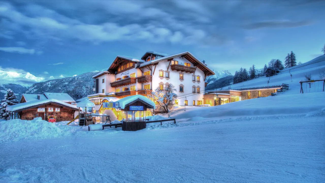 Wellness- & Familienhotel Panorama im Winter bei Dämmerung, umgeben von Schnee und Bergen, mit beleuchteten Fenstern und Skipiste.