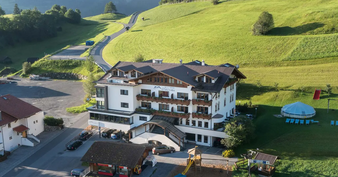 Luftaufnahme des Wellness- & Familienhotels Panorama in grüner Berglandschaft mit Spielplatz und Aussenpool.