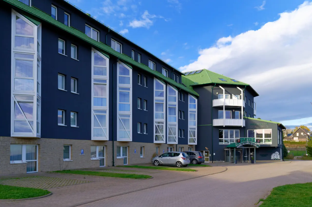 Aussenansicht des Hotel Kammweg am Rennsteig mit dunkelblauer Fassade, grünem Dach und Parkplätzen.