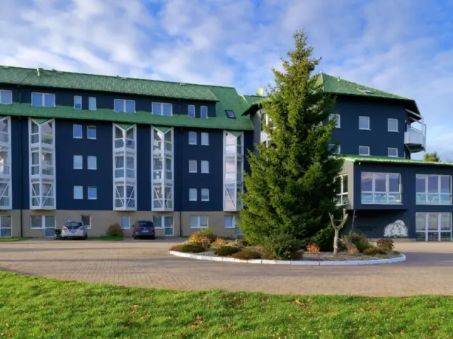 Aussenansicht des Hotel Kammweg am Rennsteig mit grünen Dächern, blauer Fassade und Vorfahrt. Ein großer Baum steht vor dem Eingang.