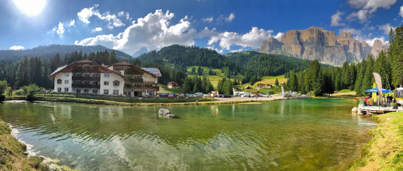 Hotel Lupo Bianco Wellness & Walking in Canazei – Panorama-Außenansicht am See mit Bergkulisse | Wellnesshotel Dolomiten