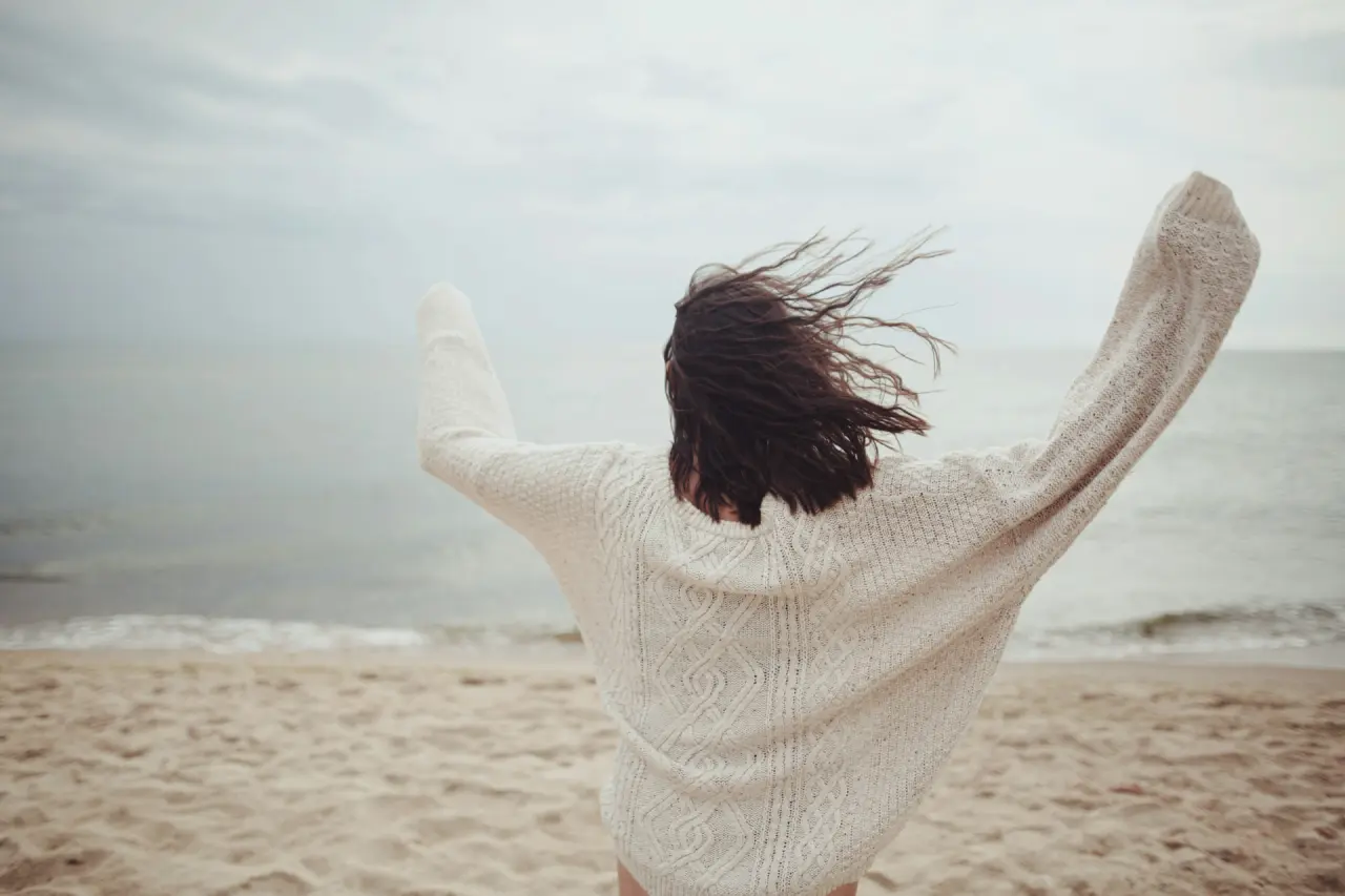 Rückenansicht einer Person mit weissem Strickpullover am Strand, die auf das ruhige Meer blickt. Entspannung am Wasser.