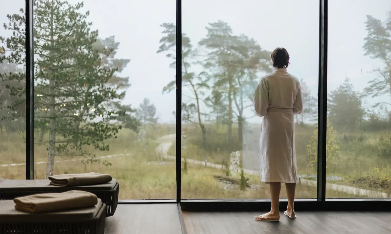 Gast im Bademantel blickt aus Panoramafenster auf Waldlandschaft im Entspannungsbereich des Hotel Sante Royale Bad Brambach.