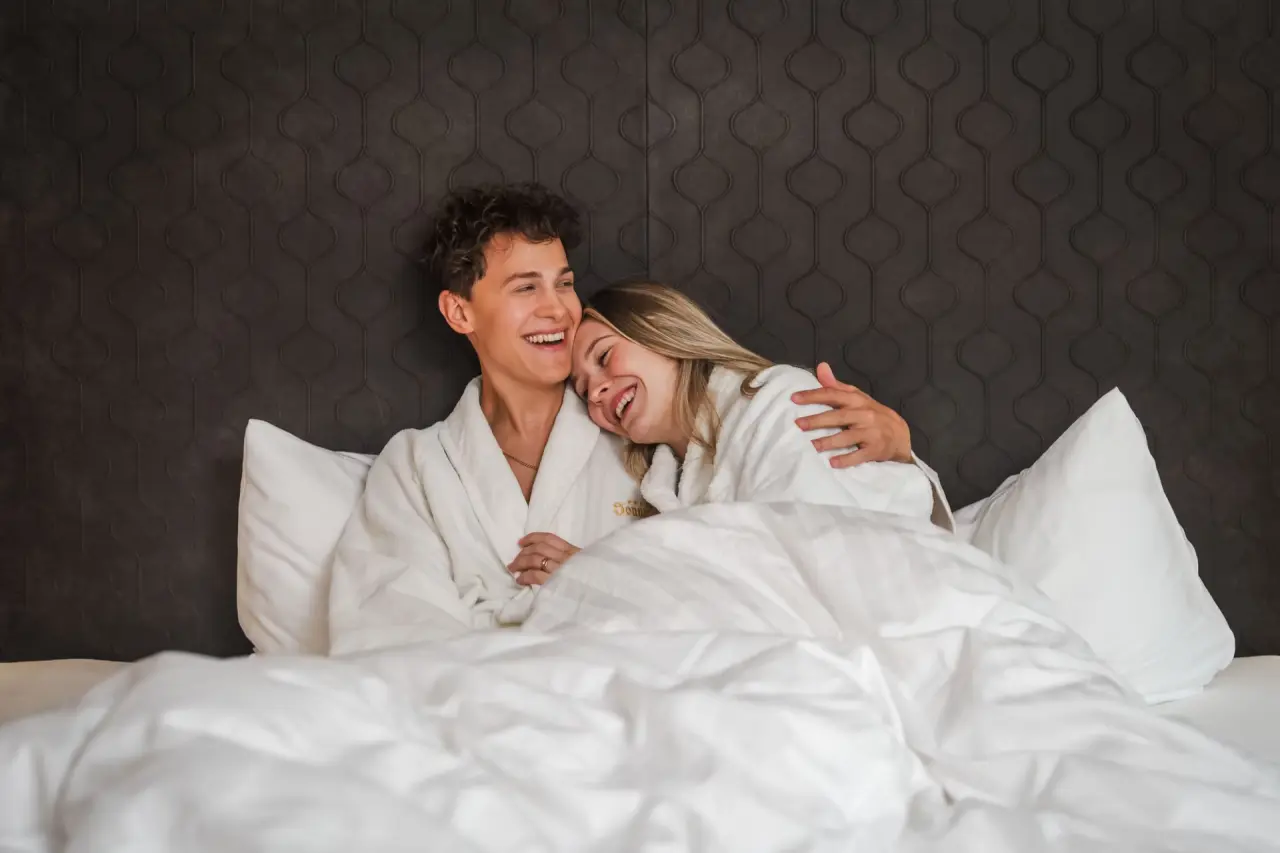 Glückliches Paar in Bademänteln entspannt im gemütlichen Hotelbett des Ferienhotel Sonnenhof****.