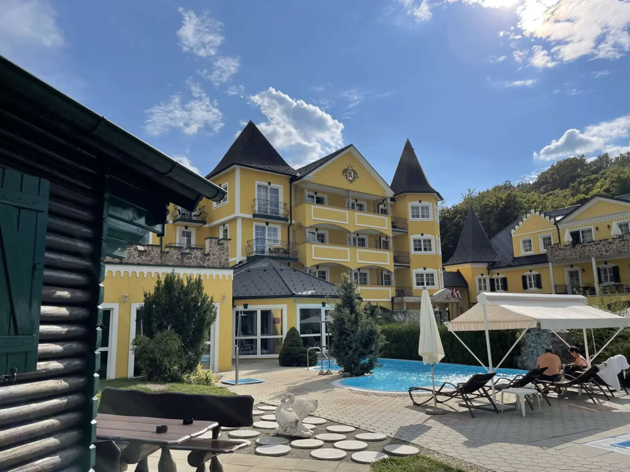 Aussenansicht des Schlössl Hotel Kindl mit gelber Fassade, Türmen und einem sonnigen Aussenpoolbereich mit Liegen.