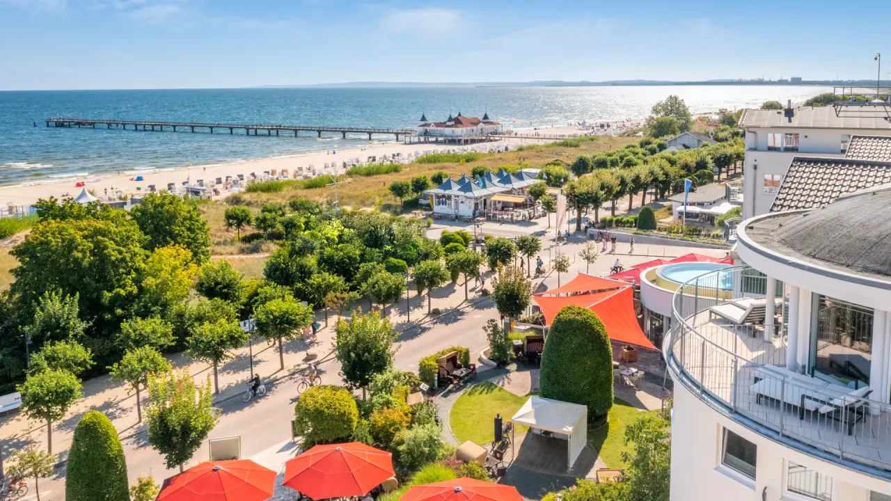 Panoramablick auf Das Ahlbeck Hotel & Spa mit Ostseestrand, Seebrücke und Hotelbalkon mit Whirlpool unter blauem Himmel.