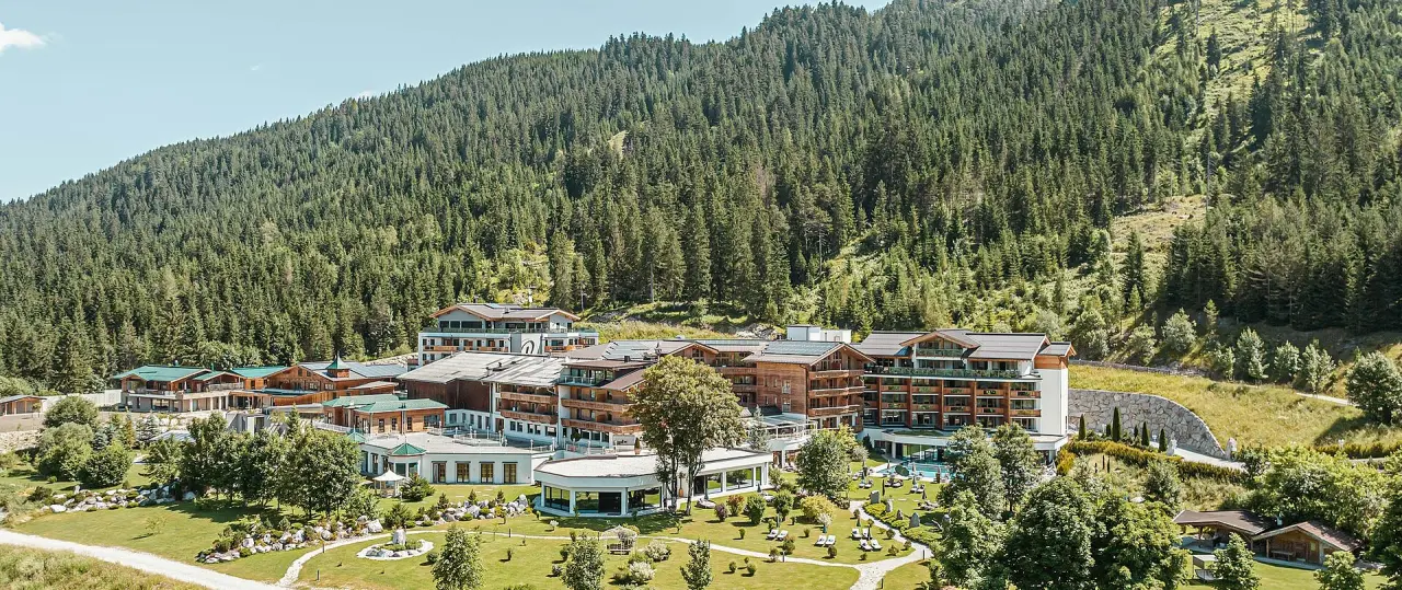 Panoramablick auf das Hotel Reiterhof in den Bergen, umgeben von dichtem Wald und einem gepflegten Garten mit Liegewiesen.