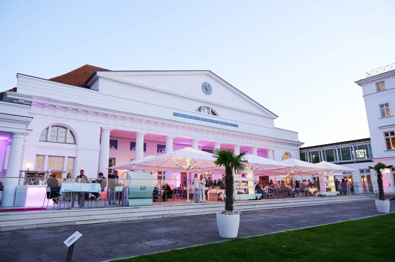 Beleuchtete Fassade des Grand Hotel Heiligendamm bei Abenddämmerung mit belebter Restaurant-Terrasse und Palmen.
