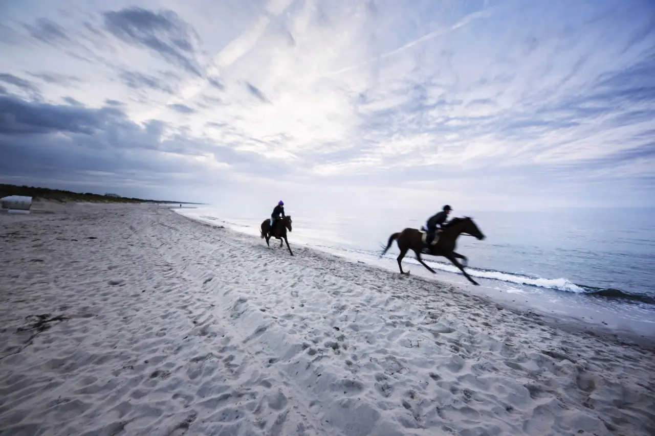 Zwei Reiter galoppieren auf Pferden am Strand des Strandhotel Dünenmeer unter bewölktem Himmel.