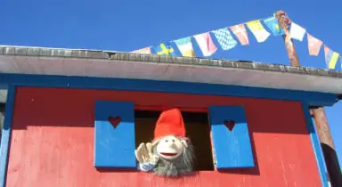 Detailansicht eines roten Gebäudes mit blauen Fensterläden und einer Puppe im Fenster, geschmückt mit bunten Fahnen, im Kleinwalsertaler Rosenhof.