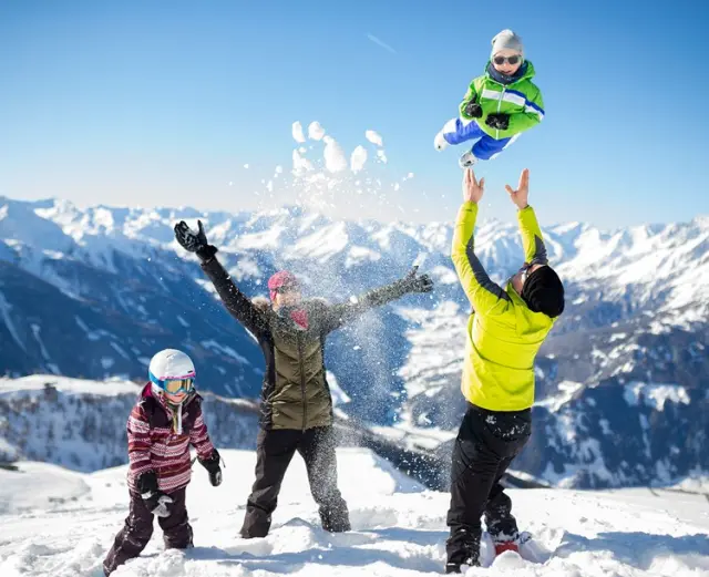 Familie spielt im Schnee mit Kind in der Luft vor verschneitem Bergpanorama im Hotel Goldried.