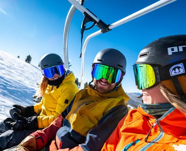 Drei Skifahrer auf einem Sessellift mit verschneiten Bergen im Hintergrund, Hotel Goldried.