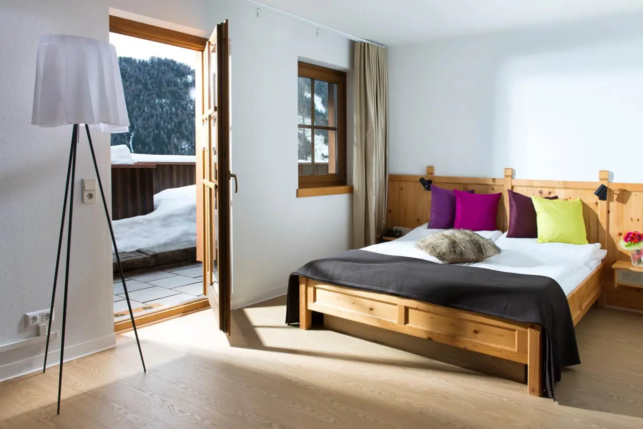 Gemütliches Doppelzimmer im Hotel Goldried mit Holzbett, bunten Kissen und Balkonblick auf verschneite Berge.