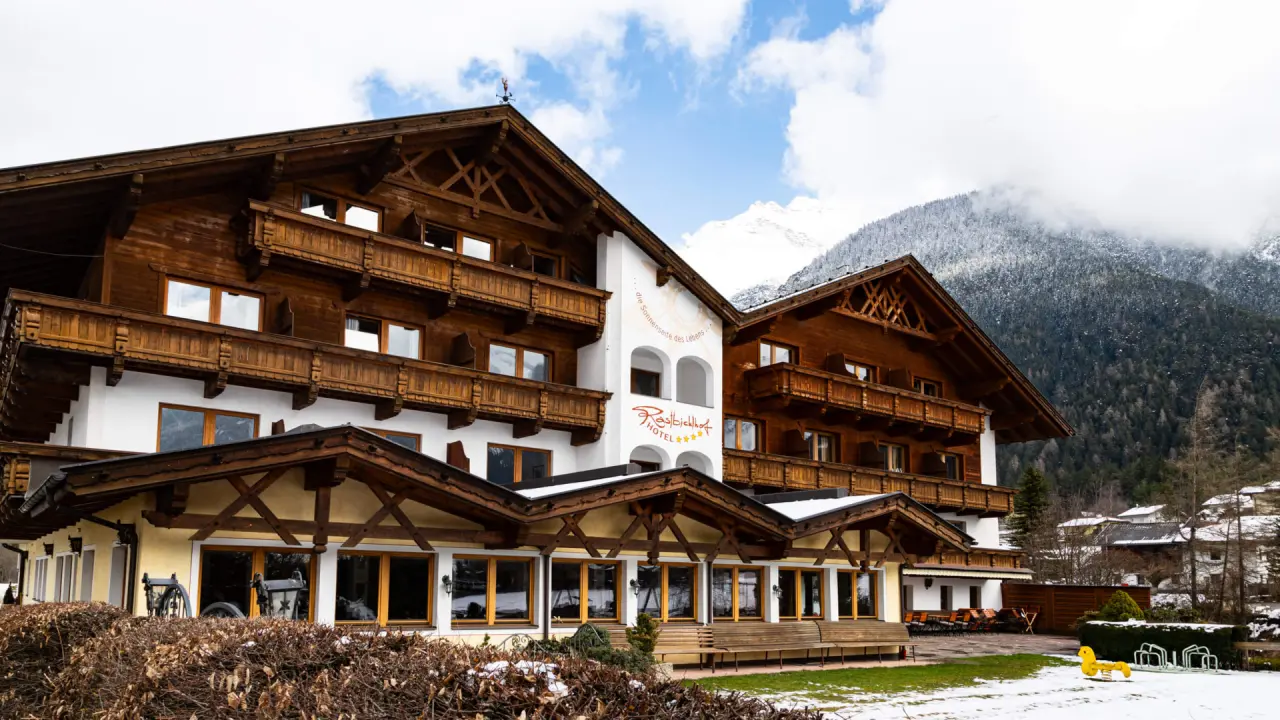 Traditionelles Hotel Rastbichlhof in den Alpen mit Holzbalkonen und schneebedeckten Bergen im Hintergrund unter blauem Himmel.