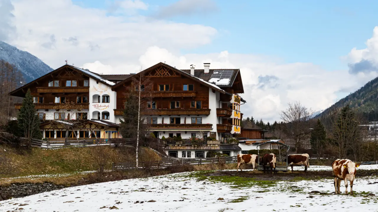Aussenansicht des traditionellen Hotel Rastbichlhof in den Alpen, umgeben von Bergen und einer schneebedeckten Wiese mit Kühen.