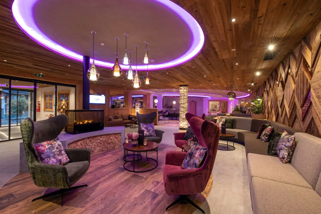 Moderne Lobby mit Kamin, gemütlichen Sesseln und lila Deckenbeleuchtung im Sportresort Alpenblick.