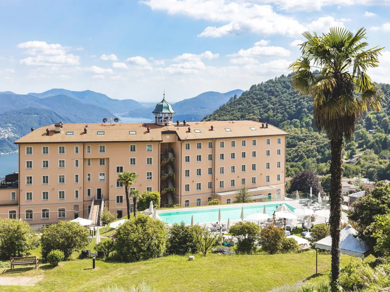 Panoramablick auf das Kurhaus Cademario Hotel mit Aussenpool, umgeben von Bergen und dem Luganersee.