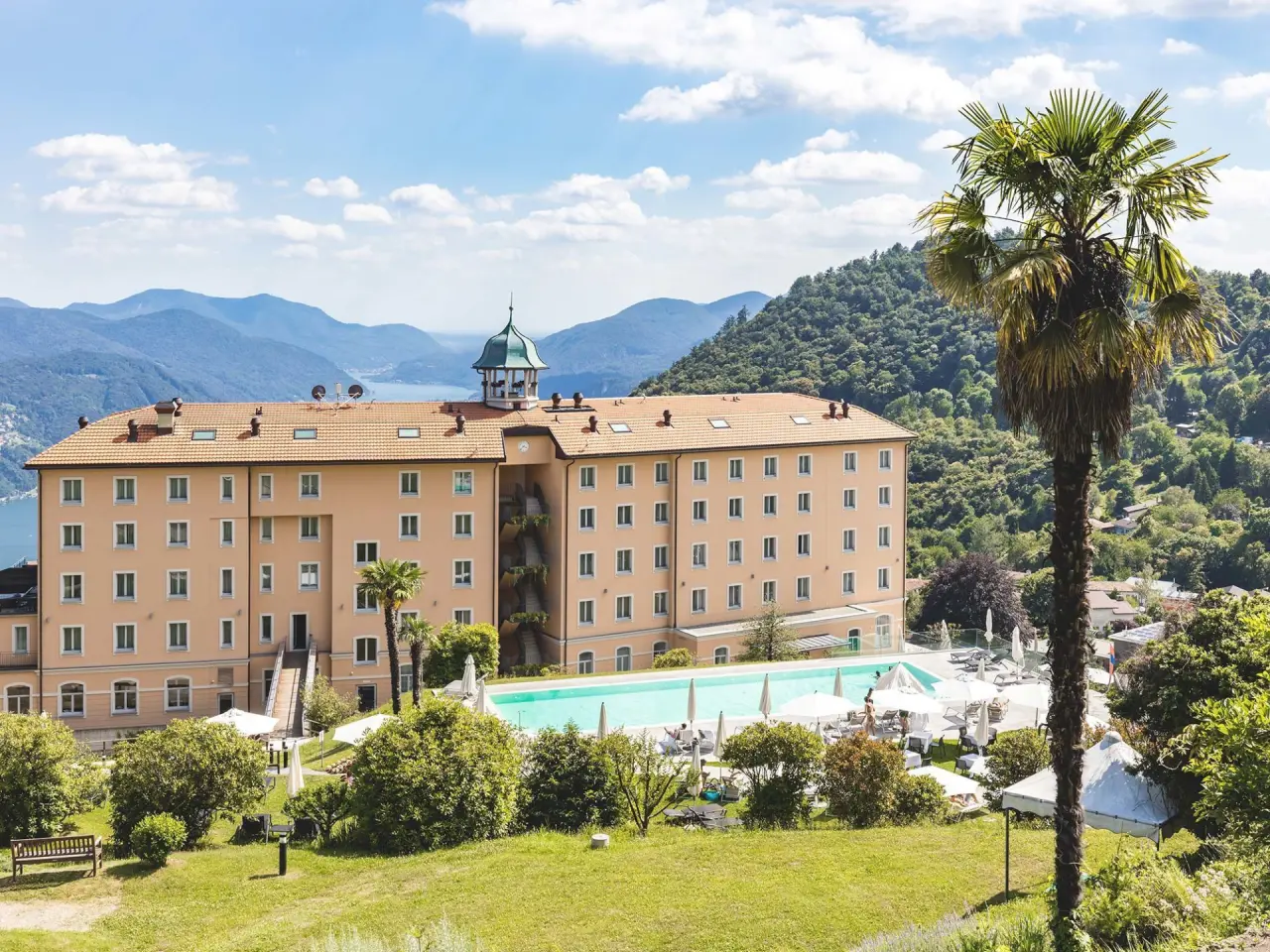 Panoramablick auf das Kurhaus Cademario Hotel mit Aussenpool, Sonnenterrasse und dem Luganersee im Hintergrund.