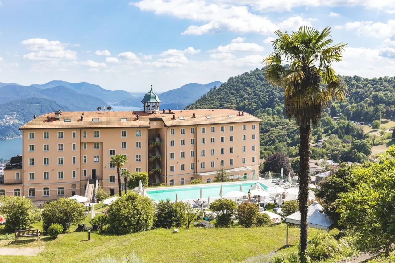 Panoramablick auf das Kurhaus Cademario Hotel mit Aussenpool, Sonnenterrasse und Luganersee im Hintergrund.