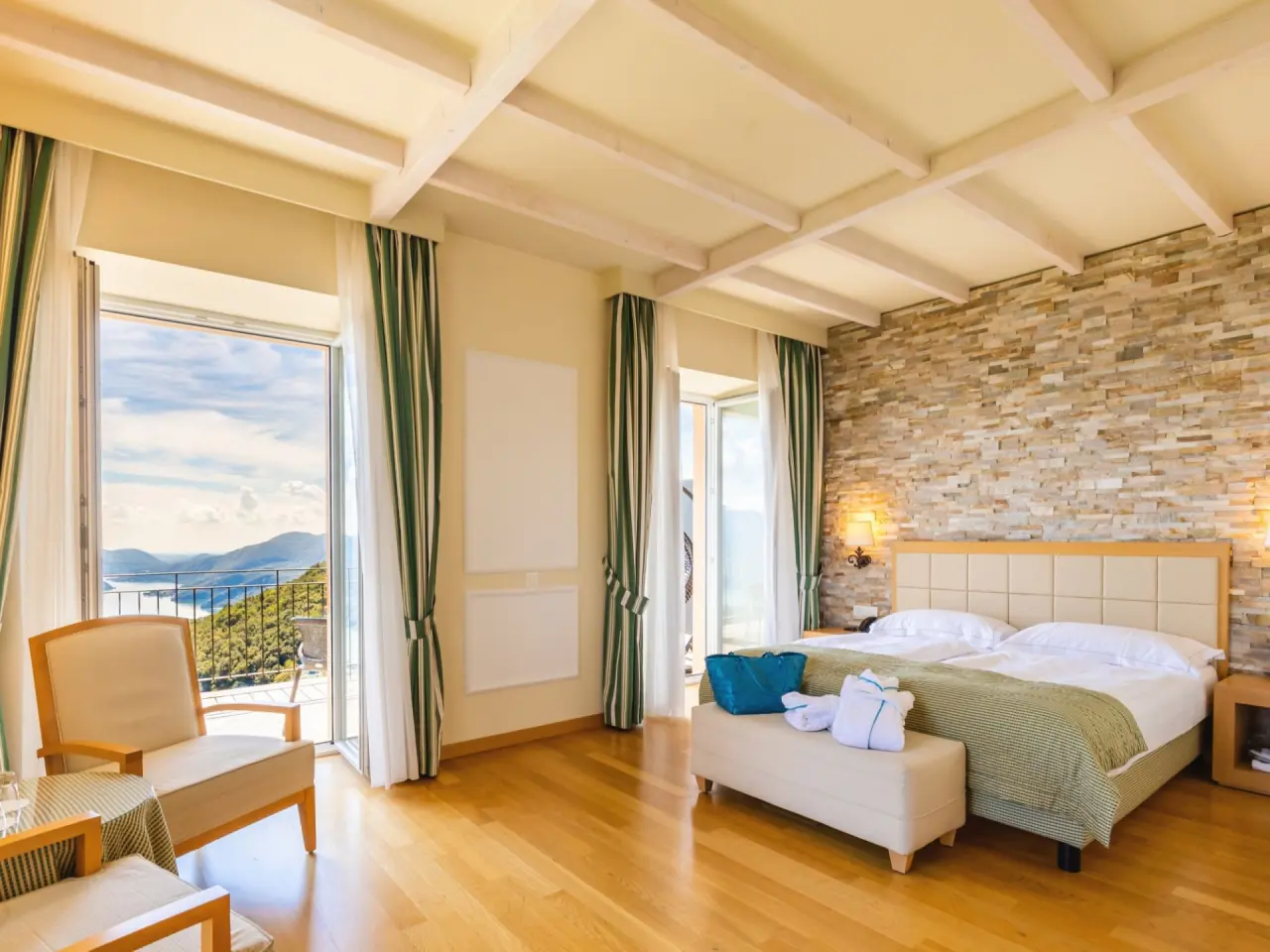 Geräumiges Hotelzimmer mit Doppelbett, Steinwand und Balkon mit Seeblick im Kurhaus Cademario.