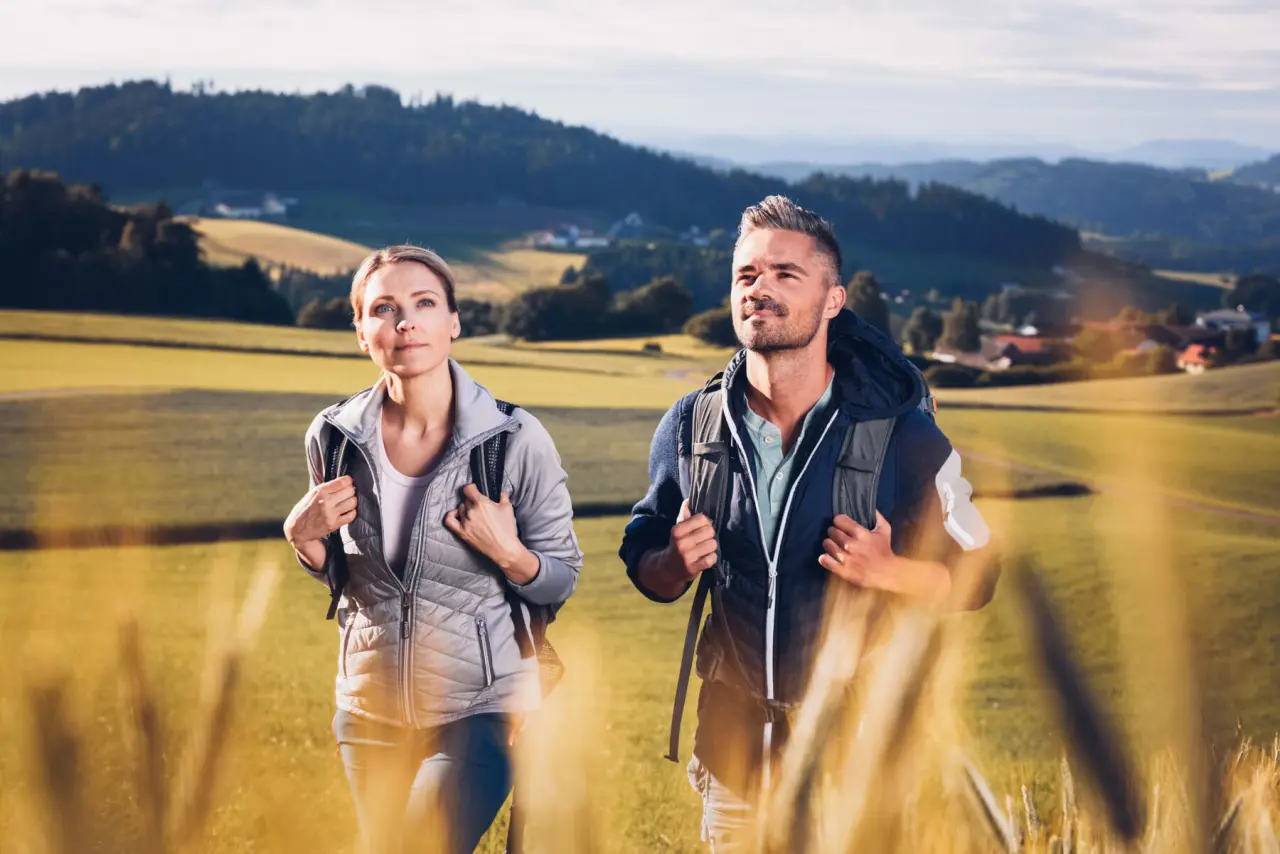 Wanderndes Paar mit Rucksäcken in sonniger Hügellandschaft nahe Hotel Lebensquell. Perfekt für aktiven Wellnessurlaub.