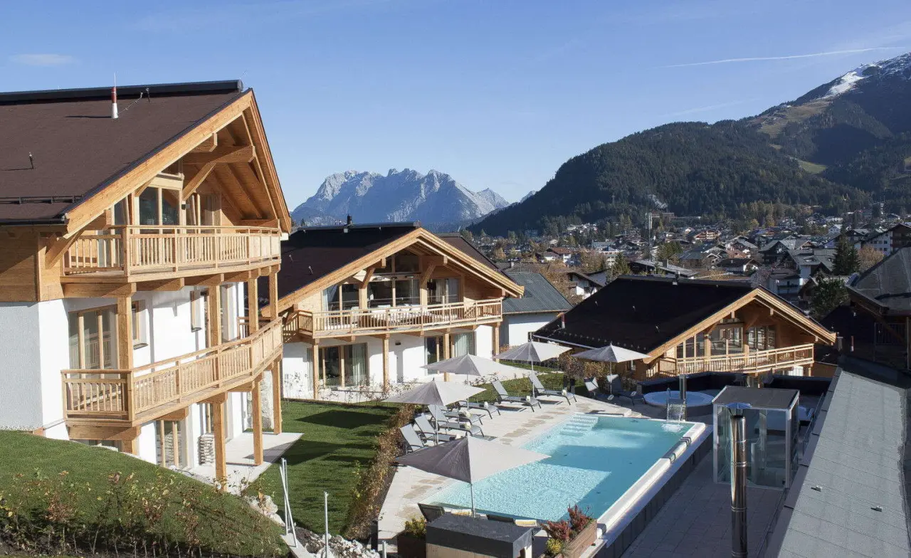 Aussenpool des Mountains Hotel mit Liegen und Sonnenschirmen vor beeindruckendem Alpenpanorama und traditioneller Architektur.