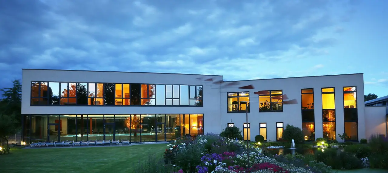 Abendansicht des See Park Janssen mit beleuchtetem Innenpool, Fitnessbereich und blühendem Garten mit Teich.