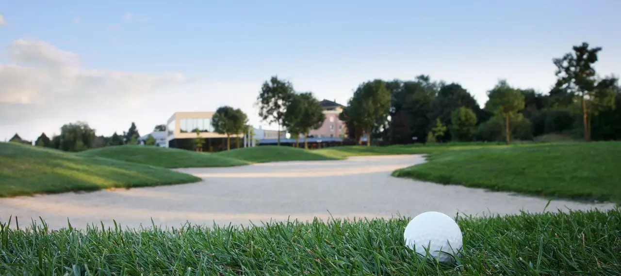 Golfball auf dem Grün mit Sandbunker und dem See Park Janssen Hotel im unscharfen Hintergrund.