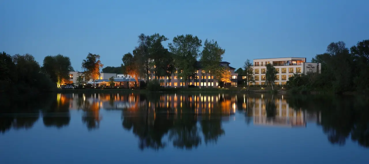 See Park Janssen Hotel bei Dämmerung, idyllisch am See gelegen mit warm beleuchteten Fenstern und Reflexionen im ruhigen Wasser.