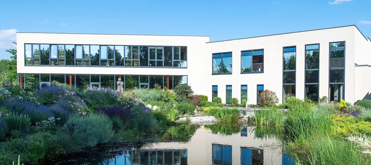 Moderne Aussenansicht des See Park Janssen Hotels mit grossen Fenstern, umgeben von einem blühenden Garten und einem Teich.