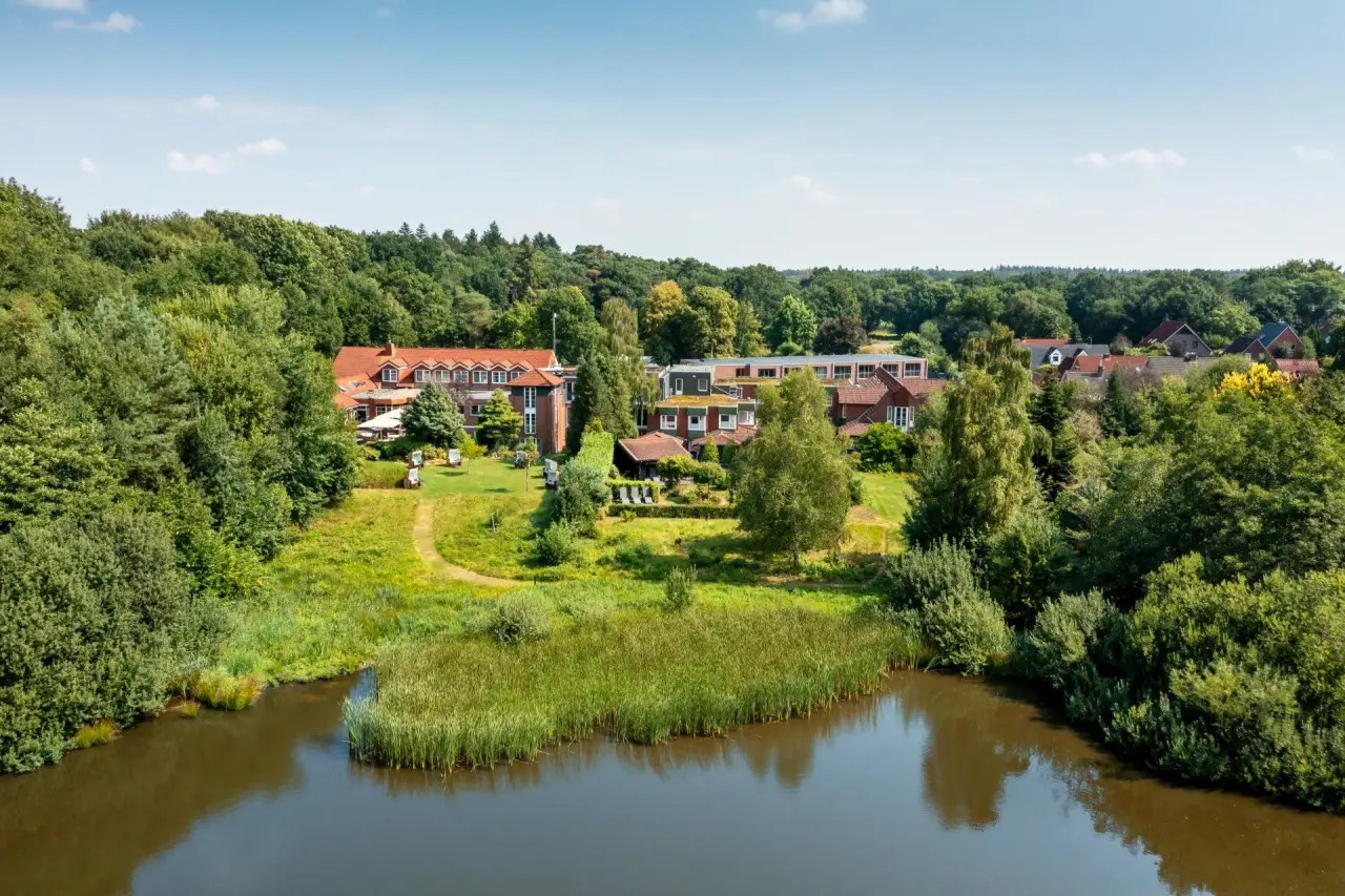 Luftaufnahme des Ringhotel Köhlers Forsthaus umgeben von Wald und einem Teich unter blauem Himmel.