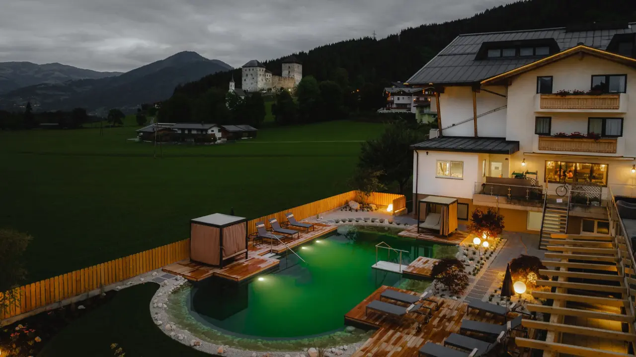 Abendliche Aussenansicht des Soulsisters’ Hotel mit beleuchtetem Naturpool und historischer Burg im Hintergrund.