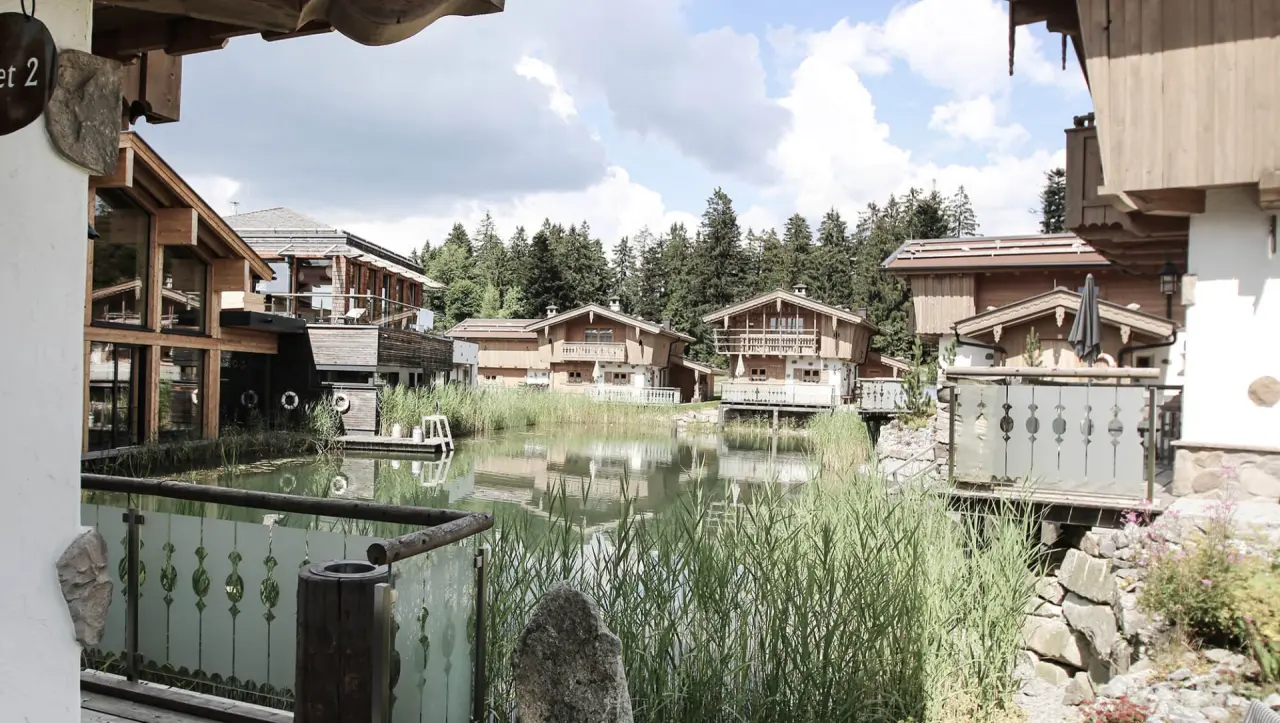 Idyllisches Chaletdorf INNs HOLZ mit traditionellen Holzhäusern am Naturteich, umgeben von Wald und Bergen.