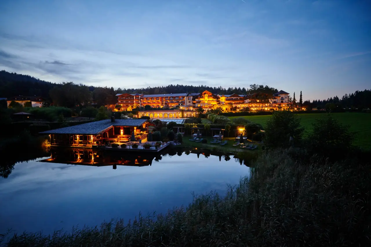 Beleuchtetes Hotel Bayerwaldhof bei Dämmerung mit Spiegelung im ruhigen See und umgebendem Wald. Einladende Abendstimmung.