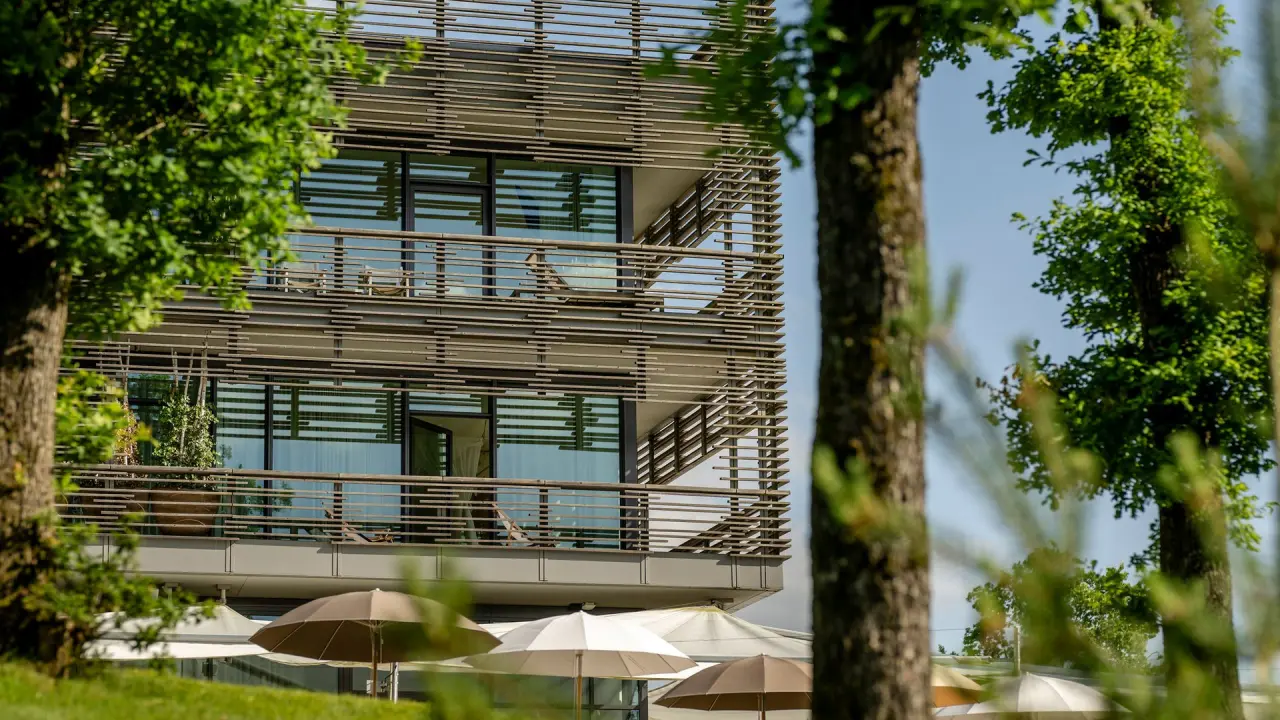 Moderne Hotelfassade des Seezeitlodge Hotels mit Balkonen, umrahmt von grünen Bäumen und Sonnenschirmen im Vordergrund.