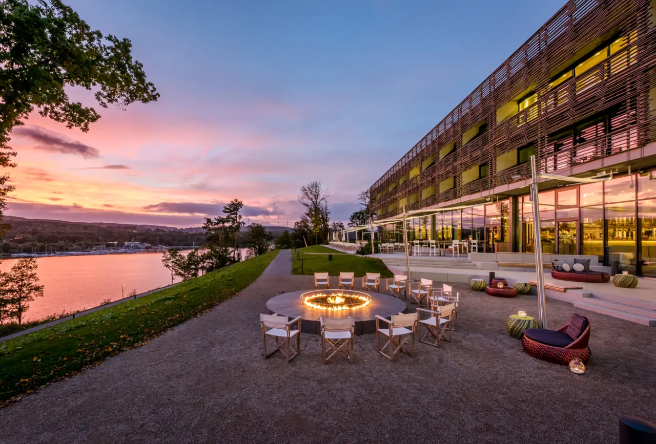 Abendstimmung am Seezeitlodge Hotel & Spa mit beleuchteter Terrasse, Feuerstelle und Blick auf den See bei Sonnenuntergang.