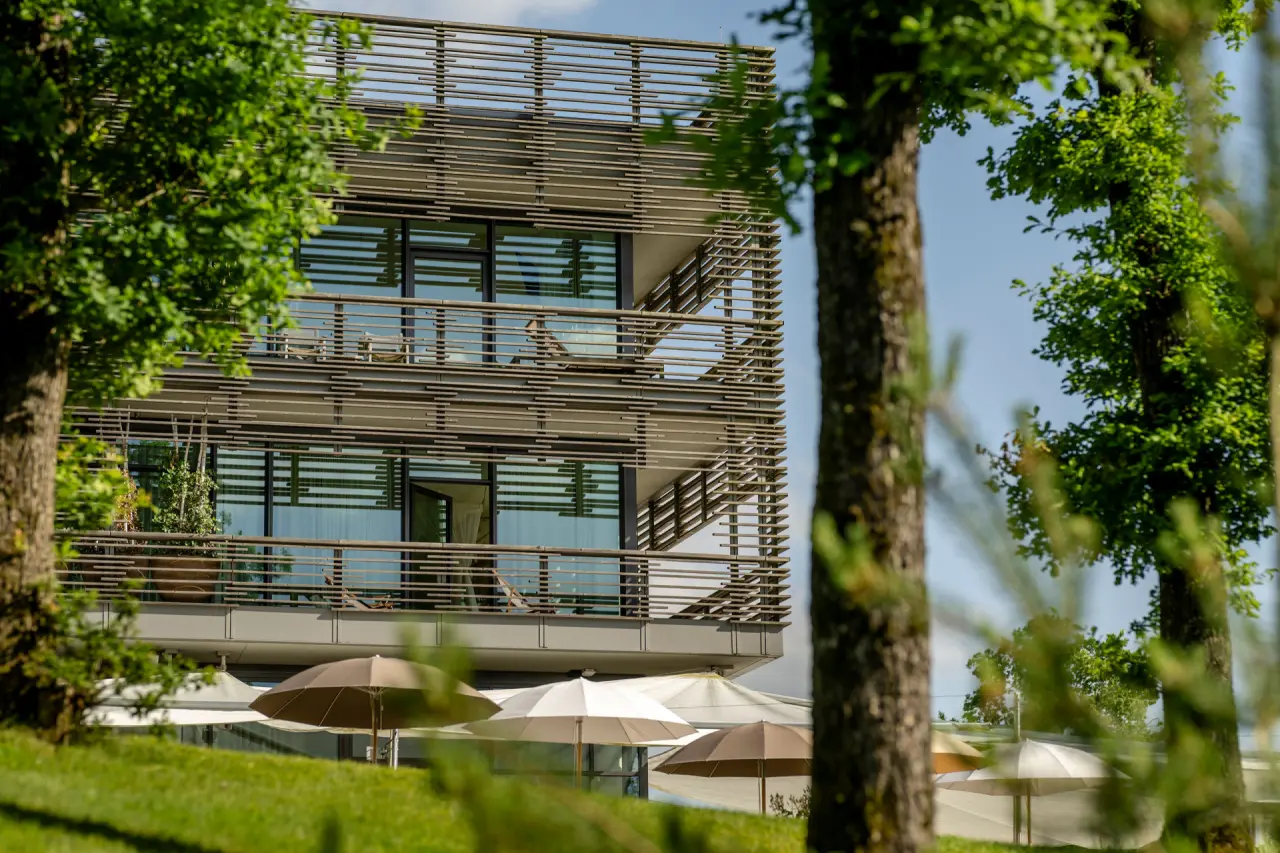 Moderne Architektur des Seezeitlodge Hotels mit Balkonen, umgeben von grünen Bäumen und Sonnenschirmen im Garten.